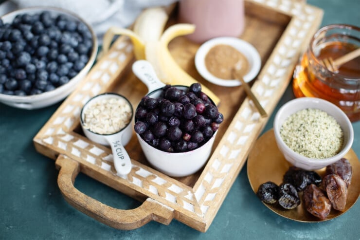 Tray with blueberries, banana, oats, almond butter, surrounded by berries, hemp seeds, honey, dates, and prunes