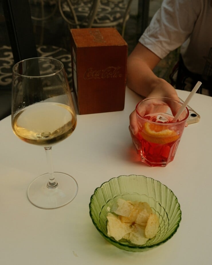 Hand holding a red cocktail at a table with a bowl of potato chips and a glass of wine