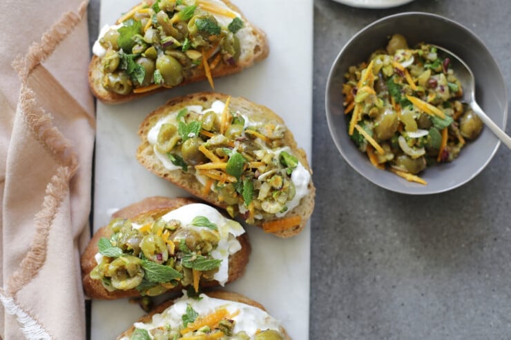 Four pieces of olive and burrata bruschetta on a serving board with olive relish on the side