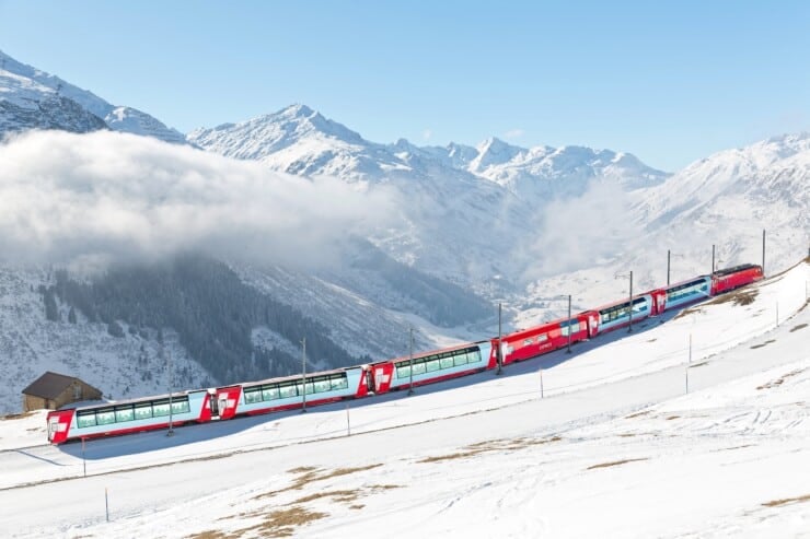 Tourists ride on a Glacier Express from St. Moritz to Zermatt on a sunny winter day and thru panoramic windows, enjoy the view of snowy mountains under blue clear sky, in Andermatt, Uri, Switzerland