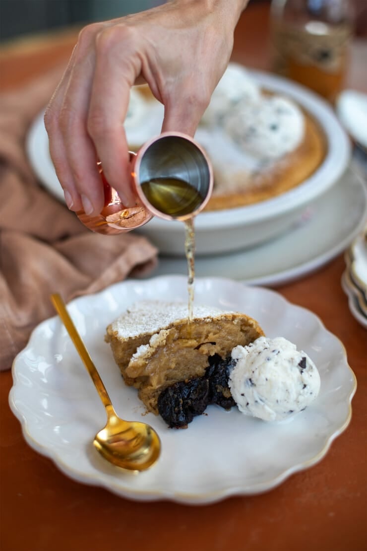 Hand with a shot glass pouring brandy over a single serving piece of prune clafoutis