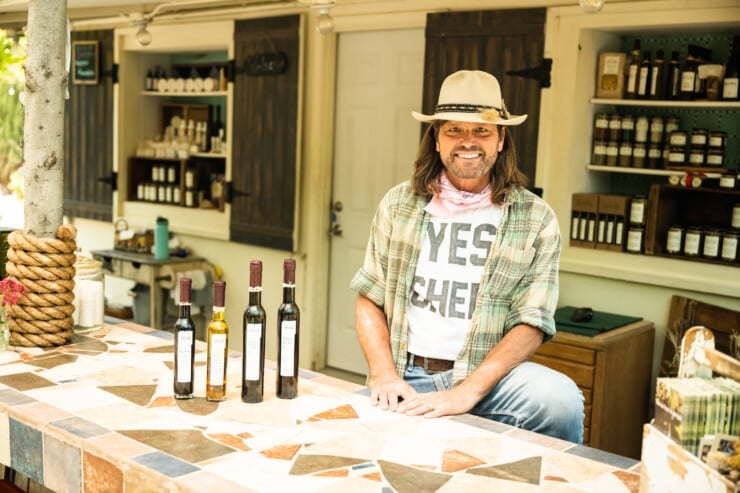 Man at a counter in jeans and a top and hat with bottles of olive oil in front of him