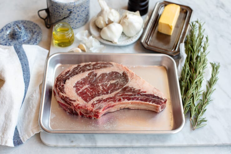 Raw bone-in ribeye on a small rimmed baking sheet surrounded by butter, rosemary, garlic, oil, salt, and pepper