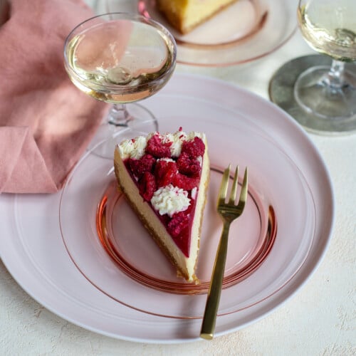 Slice of cheesecake on pink plate with fork surrounded by napkin, and glasses of wine