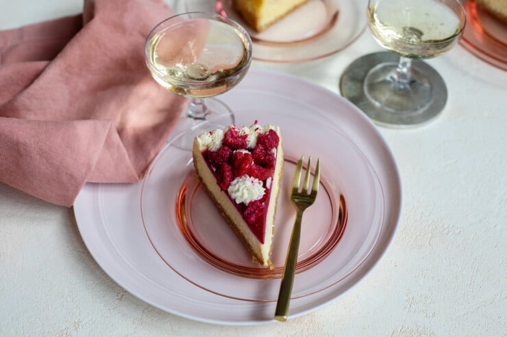 Slice of cheesecake on pink plate with fork surrounded by napkin, and glasses of wine