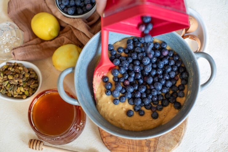 Blueberries being poured into a bowl of batter and surrounded by honey, lemons, and pistachios