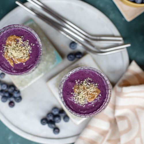 Two purple smoothies on coasters with straws, blueberries, and a napkin nearby
