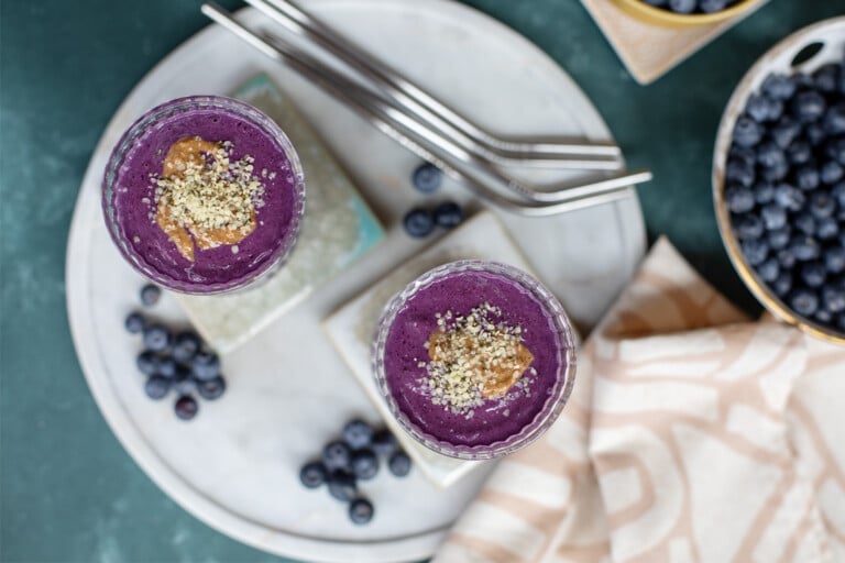 Two purple smoothies on coasters with straws, blueberries, and a napkin nearby