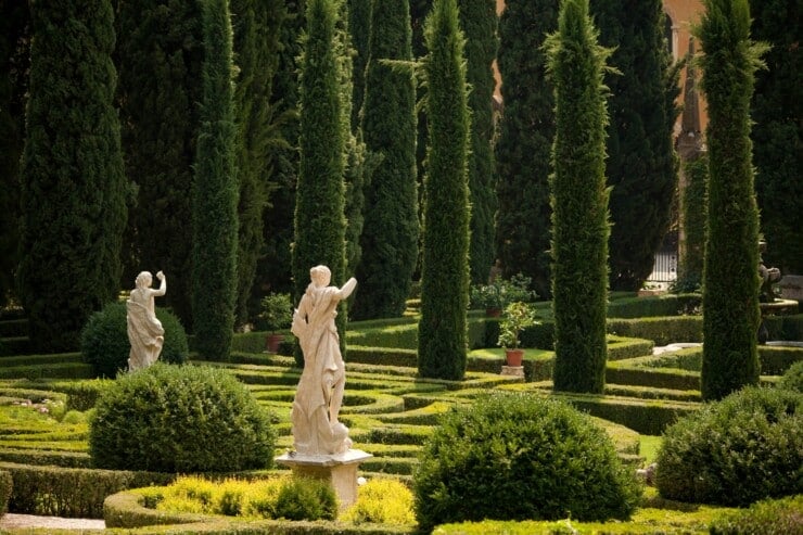 Two marble statues set among manicured hedges with Italian cypress in the background