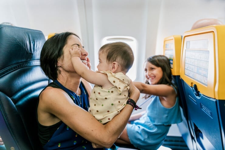 A mother shares a playful and loving moment with her baby while on an airplane, as her older child looks on and laughs. The in-flight scene captures the warmth and connection of family travel.