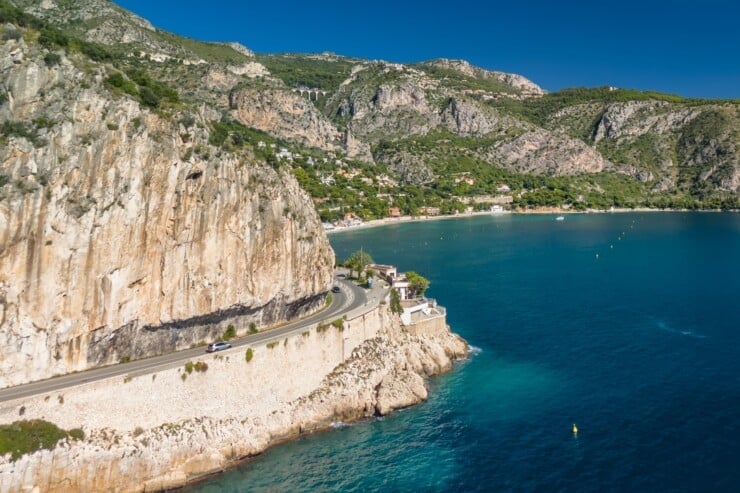 Aerial view of the French Riviera coastline between Nice and Monaco. A car drives along the Corniche Inferieure road, winding through high cliffs above the turquoise waters of the Mediterranean Sea